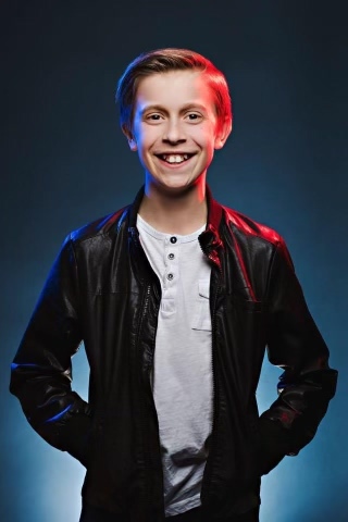 A young boy with a blue background smiling into the camera, wearing a white shirt and black jacket.