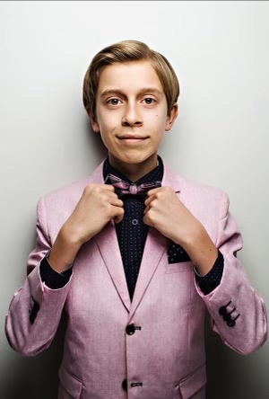 A young boy in a pink suit adjusts his bowtie while looking into the camera with a smirk.