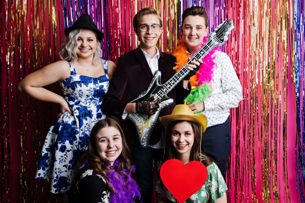 Five teens smiling into the camera while decorated with props such as a guitar, heart sign, cowboy hat, and boa.