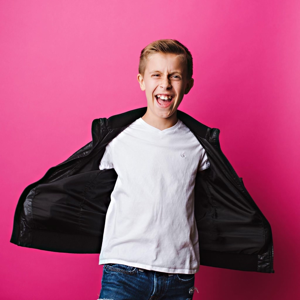 A young boy with a bright pink background, laughing at the camera with his hands in his pockets and opening his jacket. He is wearing a black jacket and white top.