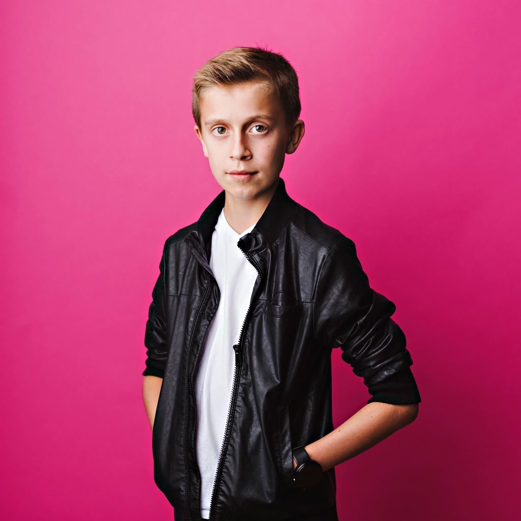 A young boy with a bright pink background, looking into the camera with his hands in his pockets. He is wearing a black jacket and white top.