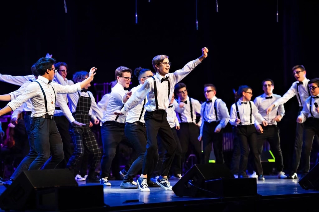 Group of boys on stage singing and dancing in black pants, white shirts and suspenders with black glasses.