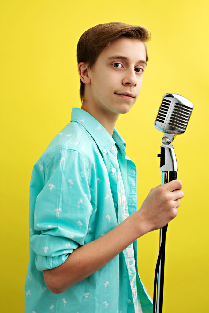 A young boy looking over his shoulder into the camera while holding a microphone. The background is bright yellow, and he is wearing a teal top.