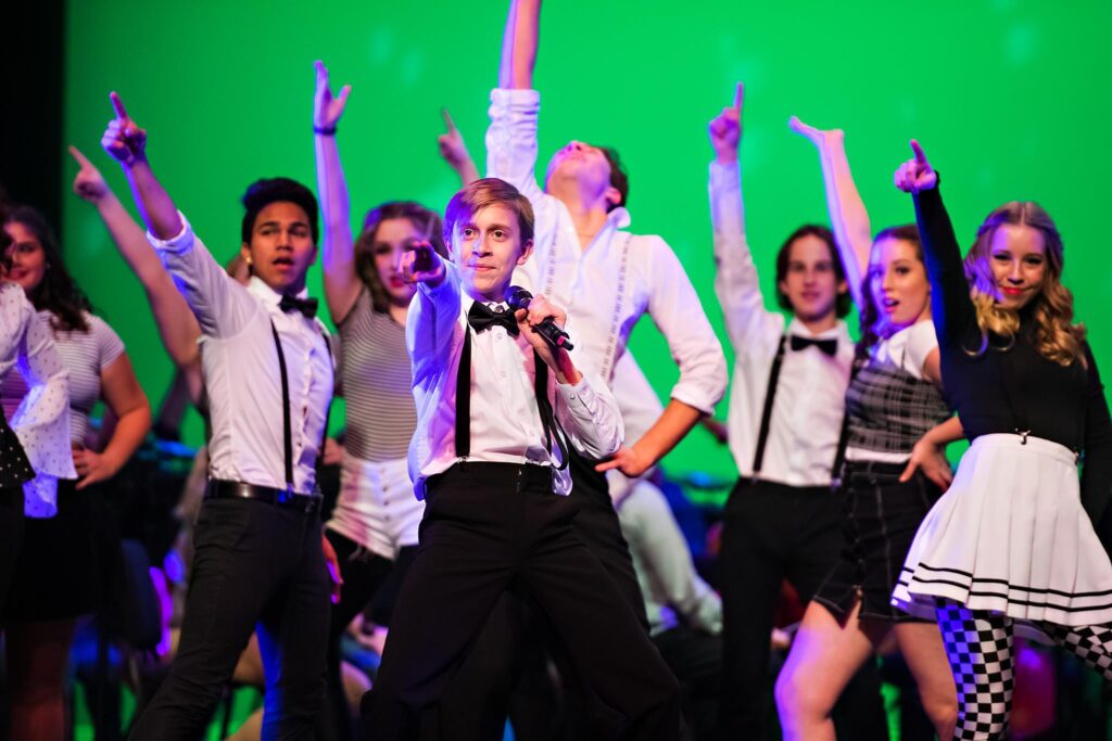 A group of young teens singing and pointing on stage wearing black and white outfits and holding microphones.