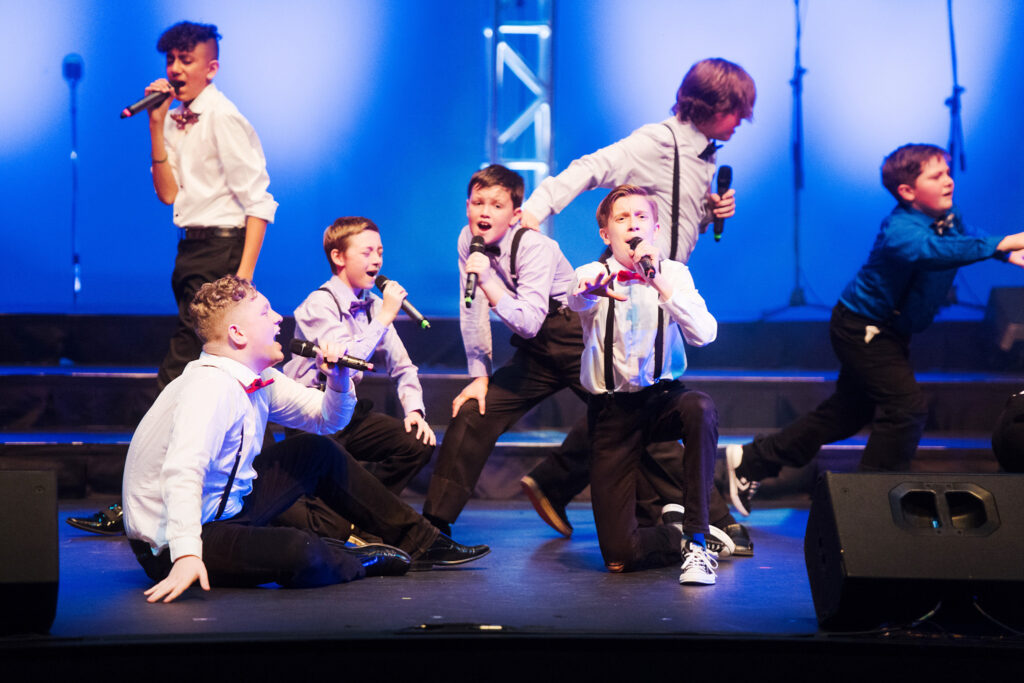 A group of boys on stage singing and dancing dramatically in black pants, white shirts, and suspenders with black glasses.