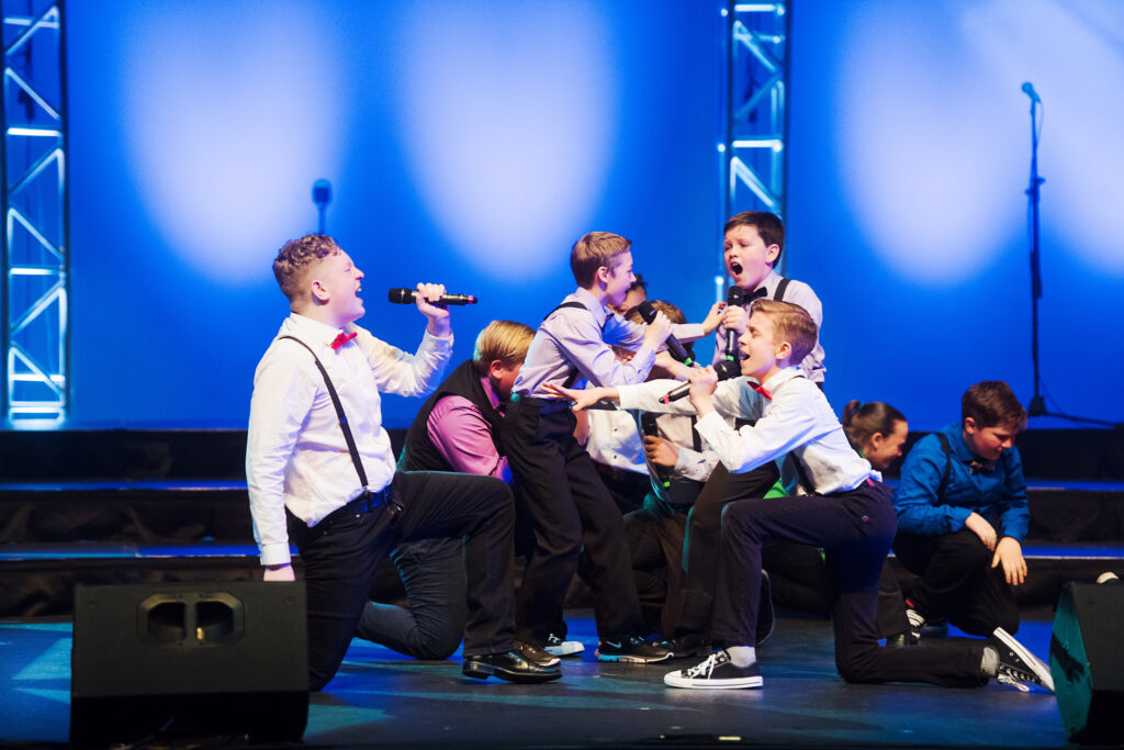 Group of boys on stage singing and dancing in black pants, white shirts and suspenders with black glasses.