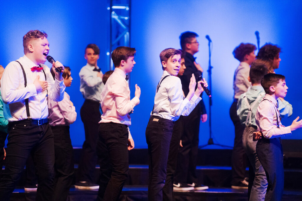 Group of boys on stage singing and dancing in black pants, white shirts and suspenders with black glasses.