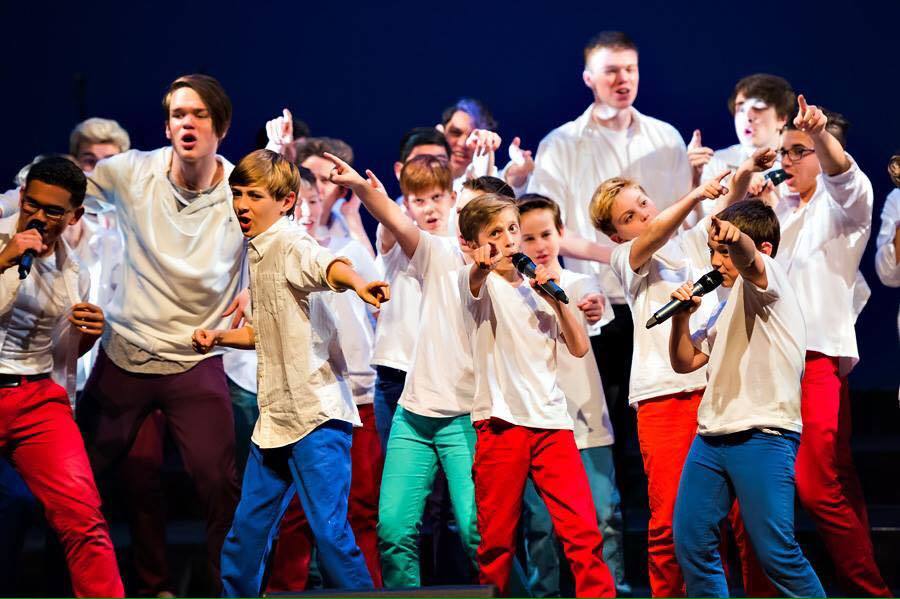 Group of boys on stage singing and pointing wearing white tops and colourful pants.