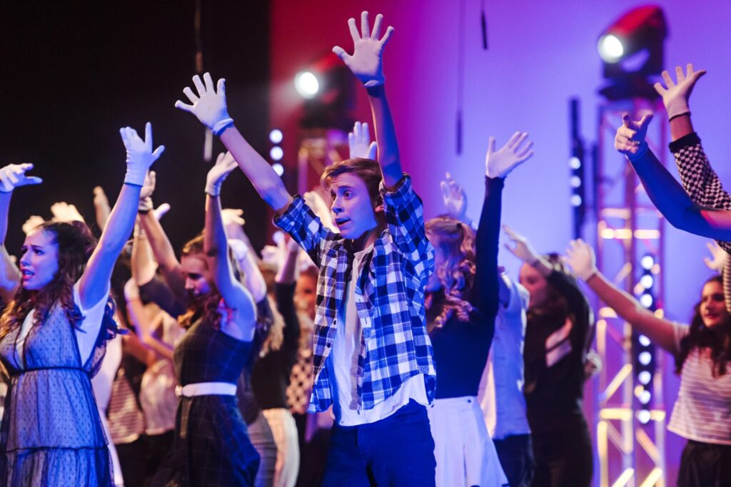 A young boy wearing flannel stands in the center of a group of people on stage, their arms raised in the air, singing.