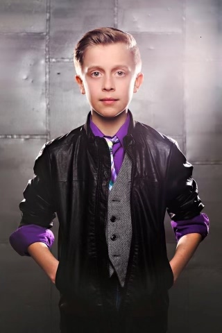 A young boy in dramatic lighting gazing into the camera, wearing a purple top and black jacket.