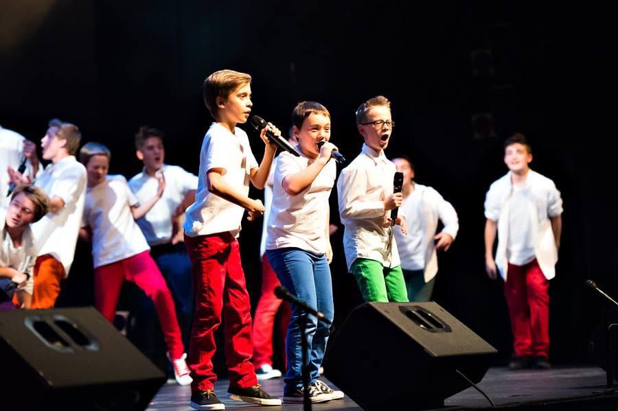 Group of soloist boys with microphones wearing white tops and colourful pants.