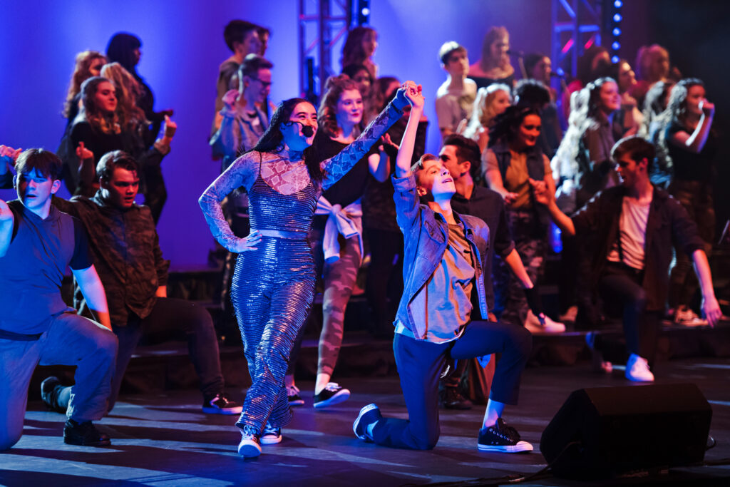 A young boy and girl are holding hands in the foreground while others are dancing on stage with purple lighting.