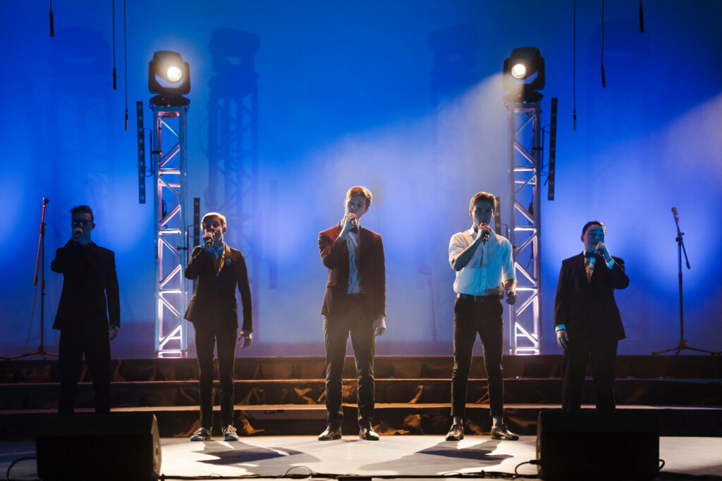 Five boys standing in a row on stage with dramatic lighting, all holding microphones and wearing suits.