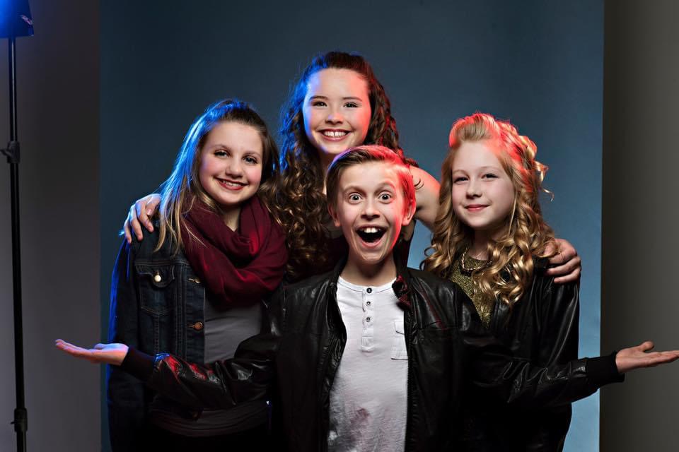 A group of three girls and one boy smiling excitedly at the camera, wearing black jackets.