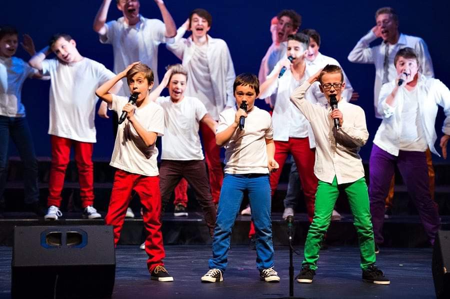 Group of boys on stage with microphones, wearing white shirts and colourful pants.