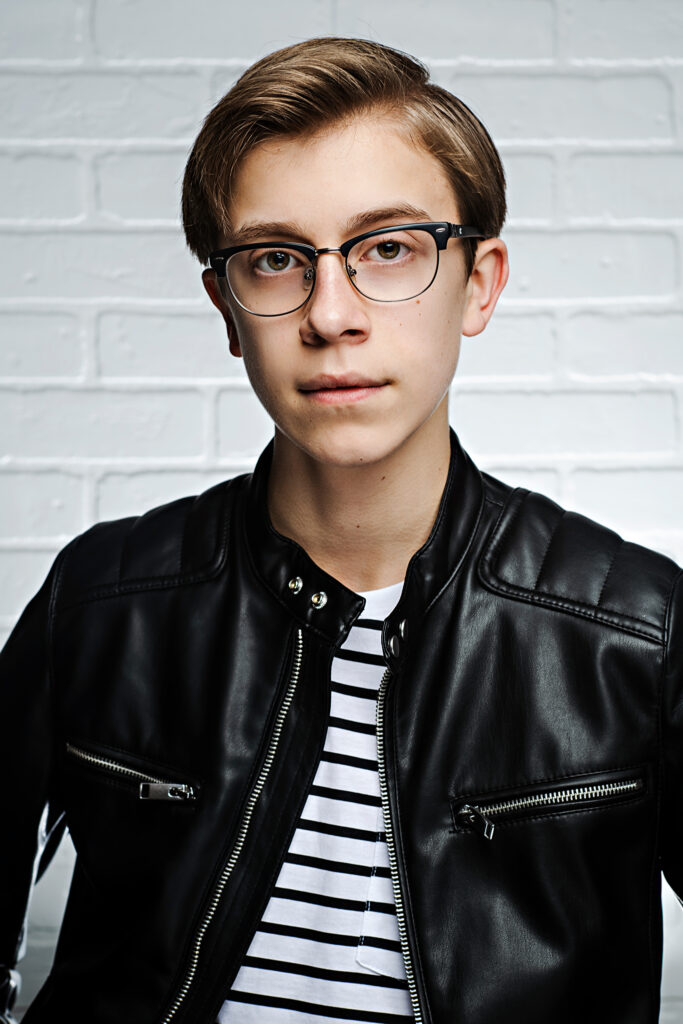 A young boy with glasses gazes into the  camera, wearing a black jacket and white and black striped top.