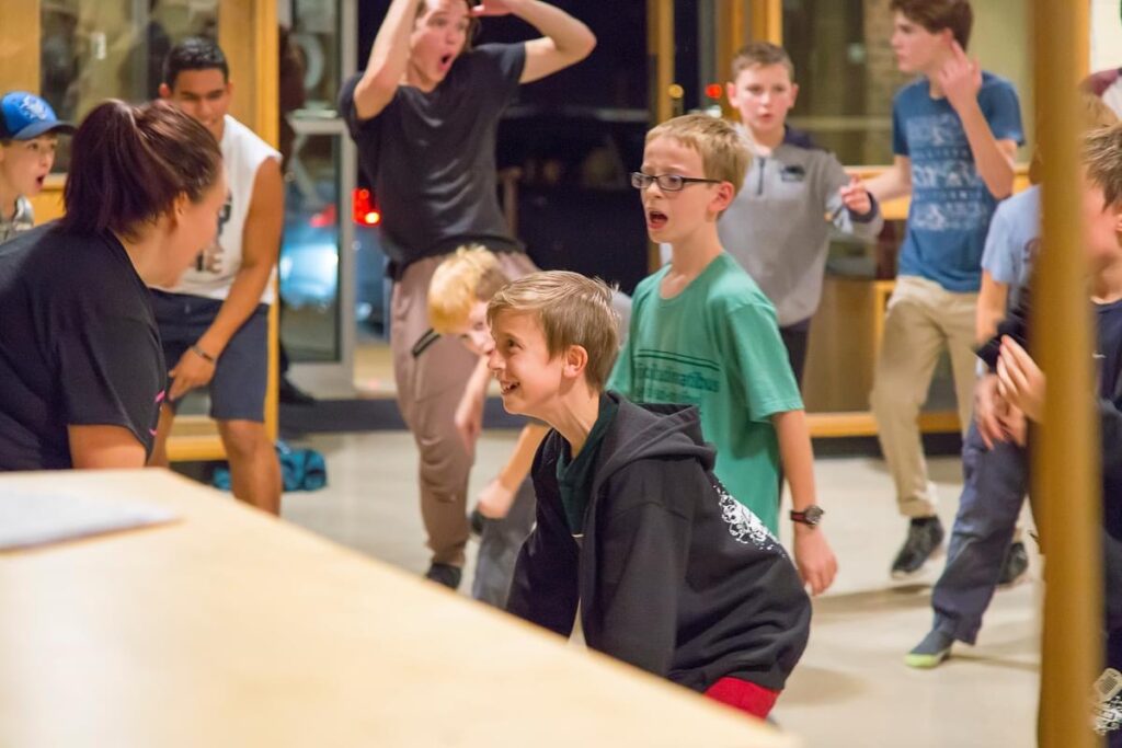 A young boy in a casual dress is squatting, learning choreography with a group of people.