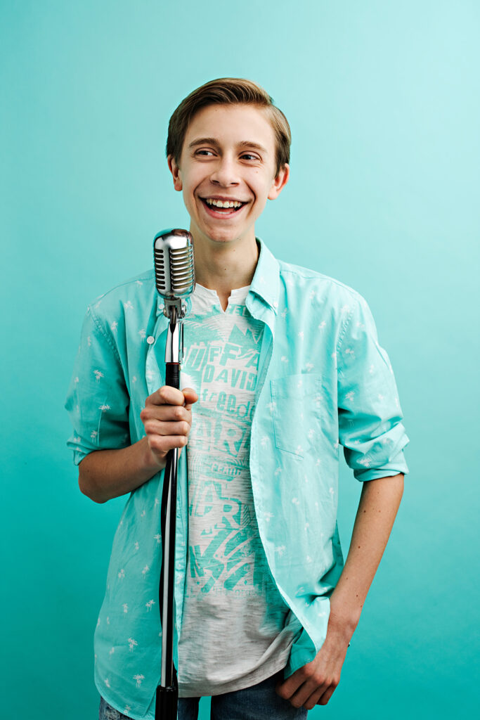 A young boy laughs as he holds a microphone, looking off-frame with a bright teal background and top.