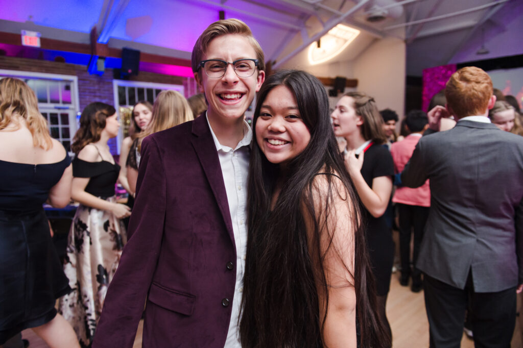 A young boy and girl standing close together and smiling into the camera, wearing formal attire. 